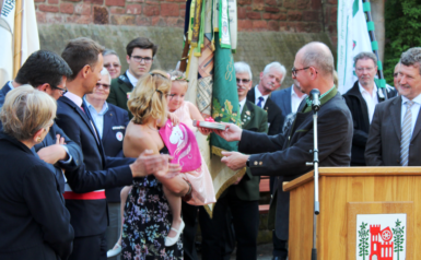 Das Bild zeigt wie das Blumen-Bärbele, das zusammen mit Bürgermeister Markus Günter das Fest eröffnet, ein Geschenk erhält. Ihr ist das Geschehen nicht ganz geheuer und kuschelt sich deshalb eng an ihre Mama. 