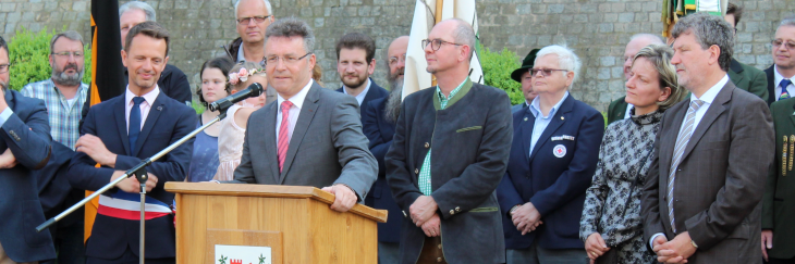 Das Bild zeigt den Landrat des Neckar-Odenwald-Kreises bei seinen Grußworten am Rednerpult. Die beiden Vertreter aus Montereau (links vom Rednerpult) und Szentgotthárd (am rechten Bildrand stehend) sind aufmerksame Zuhörer.